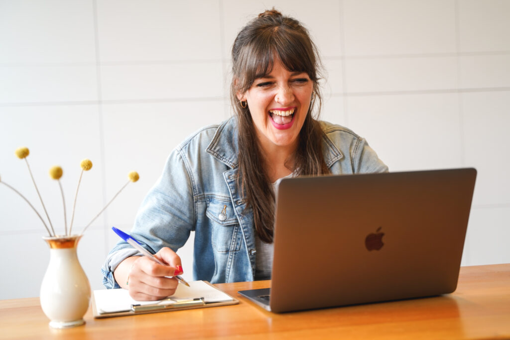 Anna sitzt vor dem Laptop und lacht herzlich. Selbstfürsorge für (soziale) Fachkräfte.